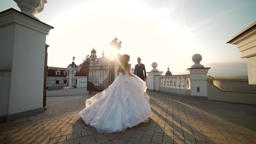 bride in a wedding dress runs to groom at sunset or sunrise against the backdrop of beautiful architecture. Slow motion. Europe. Fashion young woman goes to her husband hugs him and kisses Slow motion Royalty-Free Stock Footage #1058041609