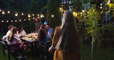 Happy multi-ethnic friends chat at garden in evening. Caucasian man gives high five to girl. African American girl sitting at table. Asian woman laughing. Friends raising and clinking glasses
