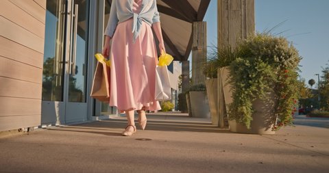Woman in pink dress with paper bags walking by shopping mall. 4K slow motion buyer shops at retail store in the outdoor shopping village on sunny summer evening at sunset. California, USA