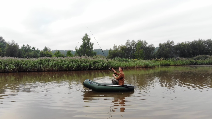 young man on an inflatable boat sits and throws a spinning or fishing rod into the water in the middle a lake or river. background of reeds and forest. Hipster guy fisherman active recreation, camping
