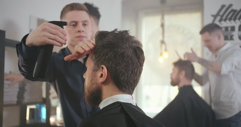 Attractive male is getting a modern haircut in barber shop. Barber wets hair by spray before cutting it. Hairdresser spraying water on hair of customers in salon