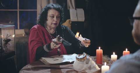 Portrait of old psychic holding snake and burning piece of paper reading spell in book with burning candles and human skull on table during seance with client