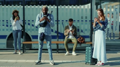 Happy Mixed-races young males and females people standing keeping safe social distance use phone on bus stop travelling street african american waiting outside vacation tourists slow motion