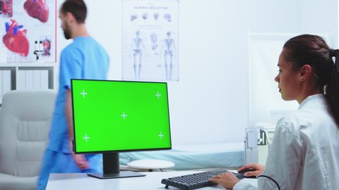 Chroma key green screen on computer in hospital cabinet while medic is working on it. Nurse in blue uniform in the background. Computer with repleceable screen used by medicine specialist in hospital – Stockvideo