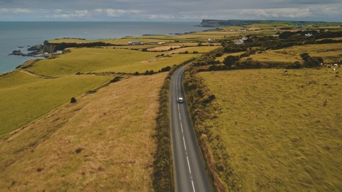 Aerial autumn road: car travel view. Beautiful Northern Ireland countryside nature scene with roadway against Atlantic cliffy shoreline in yellow fall tones. Footage shot in 4K, UHD