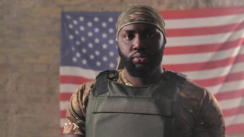 Portrait of serious african american military male in camouflage bandana and bulletproof vest posing against of USA flag. Brave bearded army male standing on background of US flag