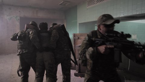 Military men helping injured comrade to leave battlefield, two military men carrying wounded soldier, others covering retreat with weapons. Squad of warriors in abandoned building insuring evacuation