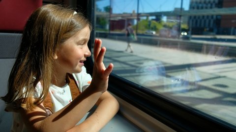 The girl travels by train and looks out the window, she waves her hand.