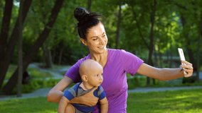 Fit woman in sportswear holding baby and using smartphone for recording video. Fitness instructor with her little child streaming for social media from park. Concept of distance training