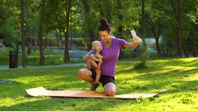 Sporty woman standing on mat and holding baby while recording video using smartphone. Fitness blogger streaming from public park together with her little child. Concept of technology
