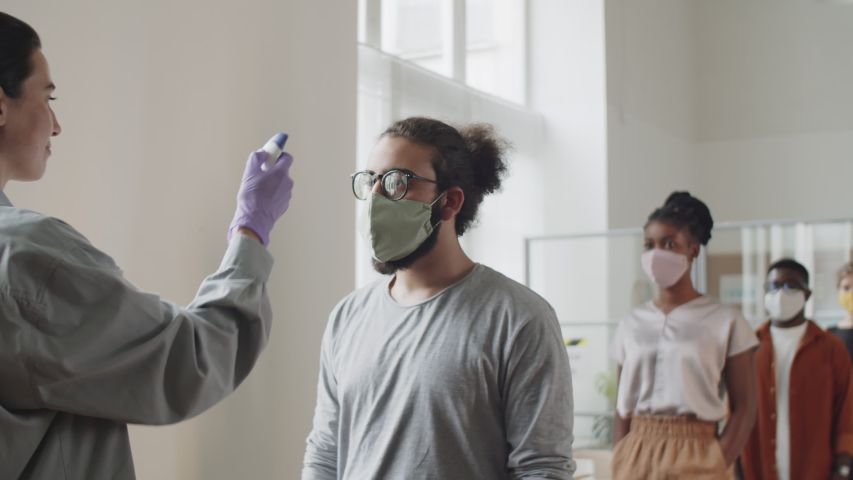 Diverse male and female colleagues in face masks walking in office and keeping social distance while manager scanning their foreheads with digital infrared thermometer during coronavirus pandemic Royalty-Free Stock Footage #1058069983