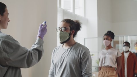 Diverse male and female colleagues in face masks walking in office and keeping social distance while manager scanning their foreheads with digital infrared thermometer during coronavirus pandemic