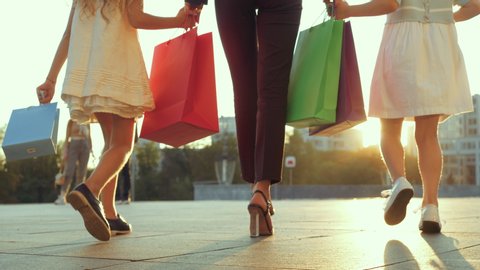 Close up of woman legs walking down street with two little girls. Mom with daughters returns from shopping, holds children by hands, carries bags of gifts shopping, modern family spends time together