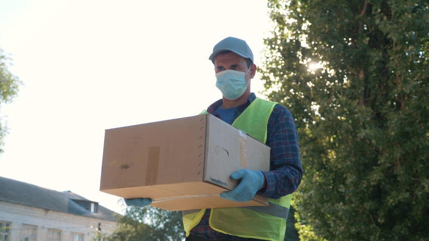 Courier wearing gloves and a mask goes to deliver food and goods during the covid coronavirus pandemic. A volunteer courier delivers online purchases. Technologies during the coronavirus pandemic Royalty-Free Stock Footage #1058072983