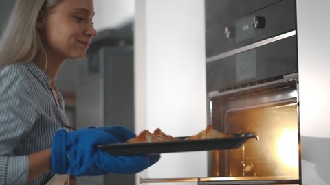 Close up of happy young woman removing hot croissants from oven at home. Pretty housewife in apron and glove taking fresh pastries from oven baking at home