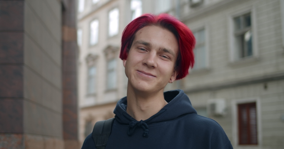 Portrait of young hipster man with red dyed hair looking to camera. Close up view of millennial guy in dark hoodie smiling while standing at street. Concept of people emotions