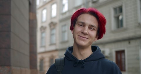 Portrait of young hipster man with red dyed hair looking to camera. Close up view of millennial guy in dark hoodie smiling while standing at street. Concept of people emotions