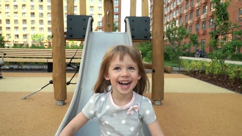 Portrait of little happy girl slides down hill, laughs and smiles on background of green summer park. Happy child plays on playground outdoors