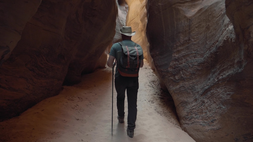 Man hiker with backpack and trekking poles hiking on bottom dry sandy cave riverbed in deep slot canyon with curved and smooth sandstone rocks walls of orange red color, slow motion, view back Royalty-Free Stock Footage #1058082349