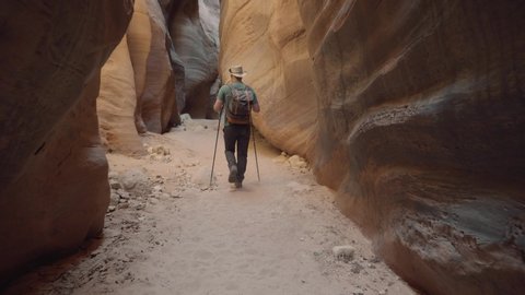 Man hiker with backpack and trekking poles hiking on bottom dry sandy cave riverbed in deep slot canyon with curved and smooth sandstone rocks walls of orange red color, slow motion, view back