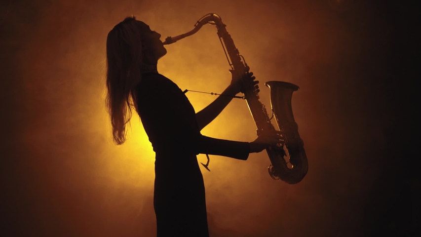 Slim young woman in smoky studio plays on saxophone, silhouette. Smoky studio