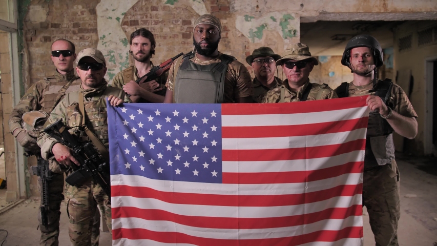 Serious multinational males soldiers in camouflage, bulletproof vests posing with US flag. Brave patriots of USA armed forces standing with weapons and holding symbol of United States of America