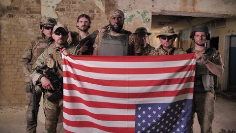 Squad of diverse US army men in camouflage holding flag upside down expressing disagreement with country's foreign policy. Multi-ethnic military males transmitting distress signal using US flag