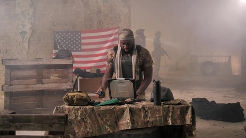 African american military man answering by walkie-talkie and working on army laptop receiving disappointing news about enemy's advance, waiting for support group at staging base of US armed forces