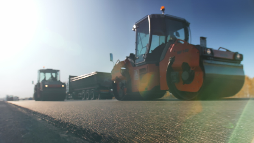A layer of freshly laid asphalt in the rays of the setting sun. Road surface repair. Construction of a new road. The rollers level and compact the asphalt. Road rollers. Sun glare. Royalty-Free Stock Footage #1058087866