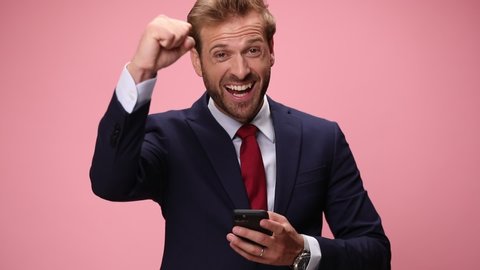 surprised elegant man in navy blue suit holding phone, reading messages, thinking, holding fist up and celebrating on pink background