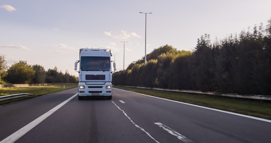Cargo truck with cargo trailer driving on a highway. White Truck delivers goods in early hours of the Morning - very low angle drive thru close up shot. Royalty-Free Stock Footage #1058088760