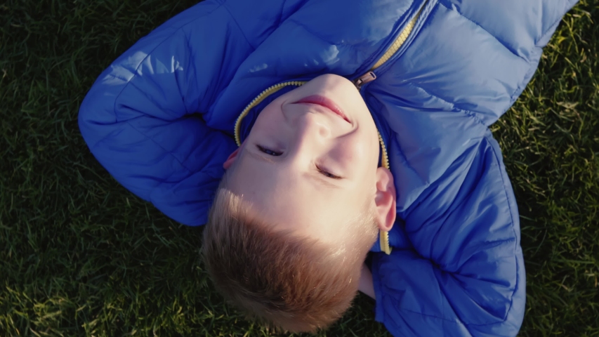 A carefree boy lies on the green grass. Smiling boy is dreaming. Dizziness. The camera is spinning. Cheerful child on the lawn near the house. View from above. Family vacation, family video. Childhood