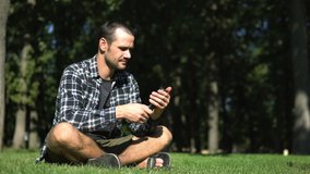 Young handsome guy in a plaid shirt leafs through the feed of social networks while sitting on the grass in the park. A young guy reads the feed of social networks on his smartphone while sitting on.