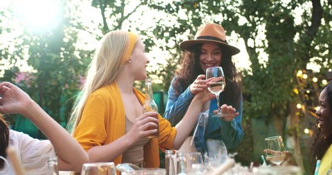 Multi-ethnic people sitting at table in garden. Caucasian female hugging African American Female. Cheerful friends spend time together. Asian woman giving high five. Raising glasses Party concept