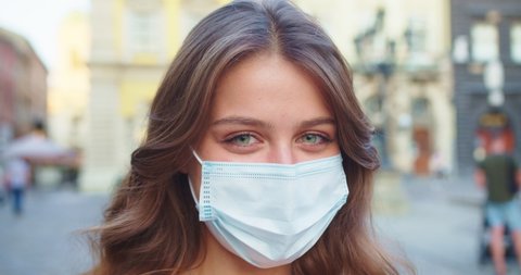 Close up portrait of joyful Caucasian beautiful young woman in medical mask standing outdoor in town. Happy smiling female putting off mask on street in quarantine. Healthcare. Safety measures concept