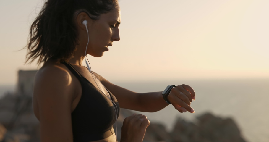 An young active sporty athlete woman with firm fit body is controlling her results after running and jogging workout and exulting satisfied of her achievement on a top of rock with seascape at sunset. | Shutterstock HD Video #1058096446