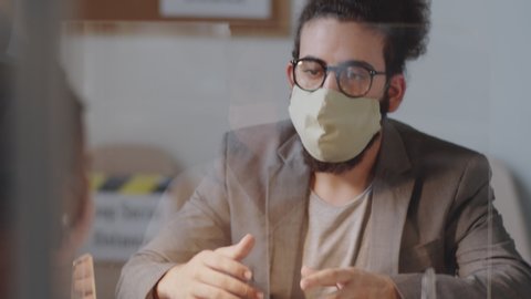 Young Arab man in face mask explaining something to afro-american female colleague through glass spit protection wall while working together in office during coronavirus pandemic