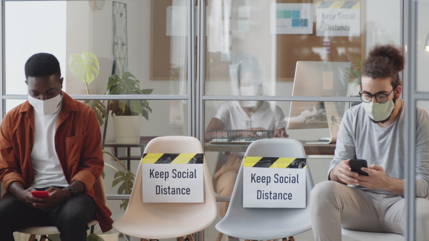 Multiethnic male coworkers sitting far apart on chairs with вЂњKeep social distanceвЂќ posters and discussing social media on smartphones while working in office during covid-19 pandemic Royalty-Free Stock Footage #1058098921