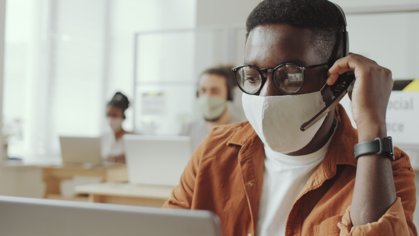 Young Afro-American man in protective face mask and headset talking on video call on laptop while working in office during coronavirus pandemic Royalty-Free Stock Footage #1058101303