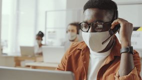 Young Afro-American man in protective face mask and headset talking on video call on laptop while working in office during coronavirus pandemic