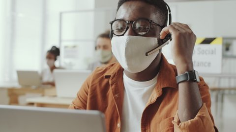 Young Afro-American man in protective face mask and headset talking on video call on laptop while working in office during coronavirus pandemic