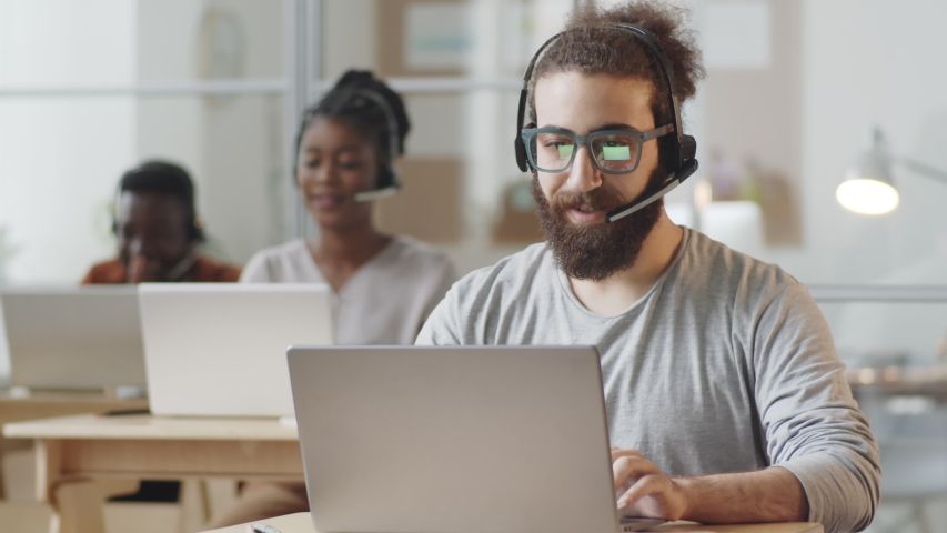 Positive young bearded man in headset smiling and typing on laptop while working in customer service center Royalty-Free Stock Footage #1058102206