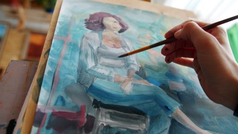 Close-up of paint-covered hand painting portrait of woman during arts class using water colors and paper on easel while student learning paint.