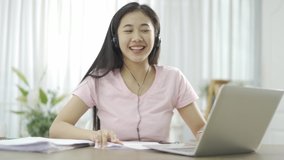 Young Asian woman wears pink shirt and black headset conference calling on laptop talks with online teacher studying, working from home. Lady student e learning using computer webcam