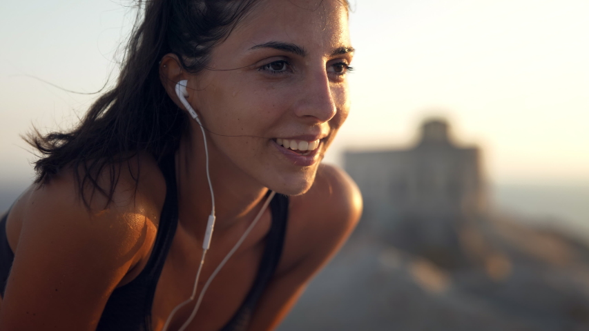 Close up of an young active sporty athlete smiling woman is taking a break after making running and jogging workout on a top of rock with seascape at sunset.