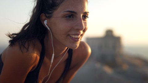 Close up of an young active sporty athlete smiling woman is taking a break after making running and jogging workout on a top of rock with seascape at sunset.