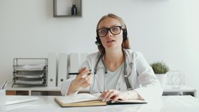 Professional female doctor in lab coat, glasses and headset sitting at table in medical office, looking at camera and taking notes while giving online consultation on video call