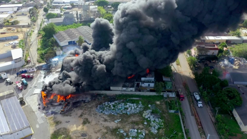 Flying Close Over a Massive Devastating Fire as it Consumes Large Commercial Warehouse Building Creating Huge Thick Black Smoke Cloud during Daytime.