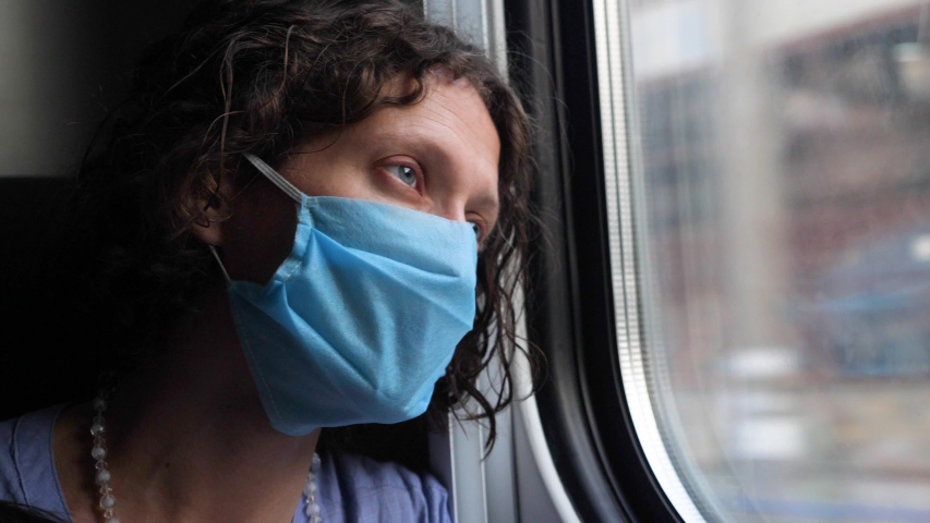 Business woman with tired eyes goes on a train trip looking out the window. Young female in a disposable medical mask watching running buildings, poor neighborhoods, coronavirus destroys the economy Royalty-Free Stock Footage #1058119177