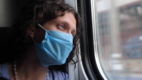 Business woman with tired eyes goes on a train trip looking out the window. Young female in a disposable medical mask watching running buildings, poor neighborhoods, coronavirus destroys the economy